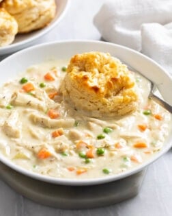 Side view of Crock Pot Chicken Pot Pie in a white bowl with a fork on the side and a biscuit on top.