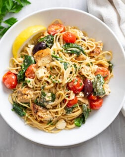 A white bowl of Mediterranean Pasta topped with tomatoes, chicken, olives, spinach, and feta with a lemon wedge on the side.