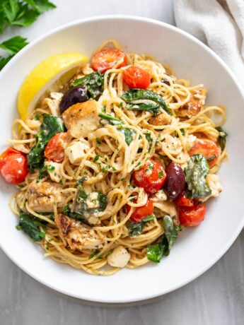 A white bowl of Mediterranean Pasta topped with tomatoes, chicken, olives, spinach, and feta with a lemon wedge on the side.