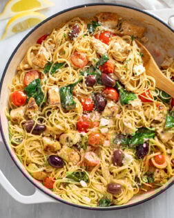 Mediterranean Pasta in a skillet with spinach, tomatoes, olives, geta, and chicken with a wooden spoon on the side.