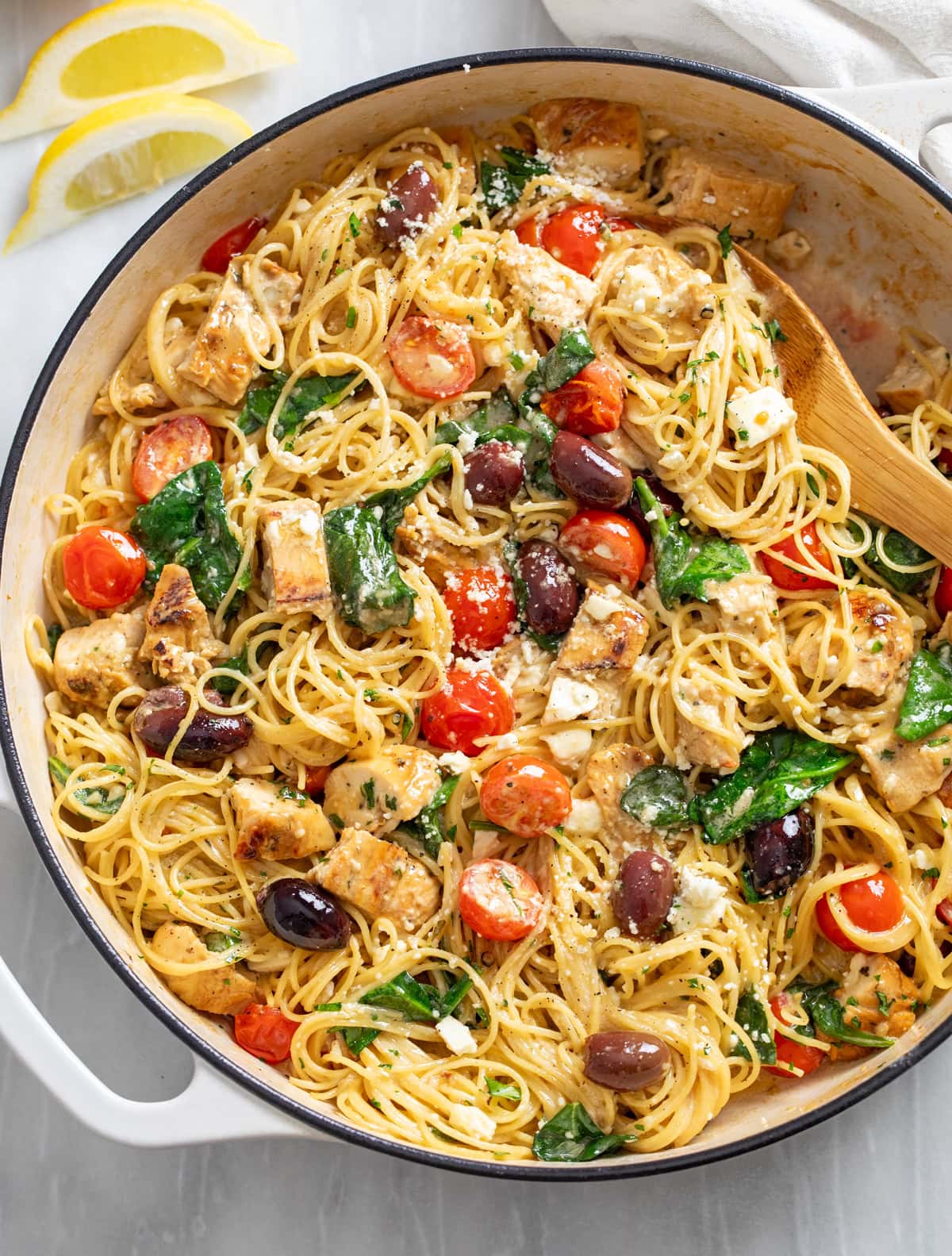 Mediterranean Pasta in a skillet with spinach, tomatoes, olives, geta, and chicken with a wooden spoon on the side.