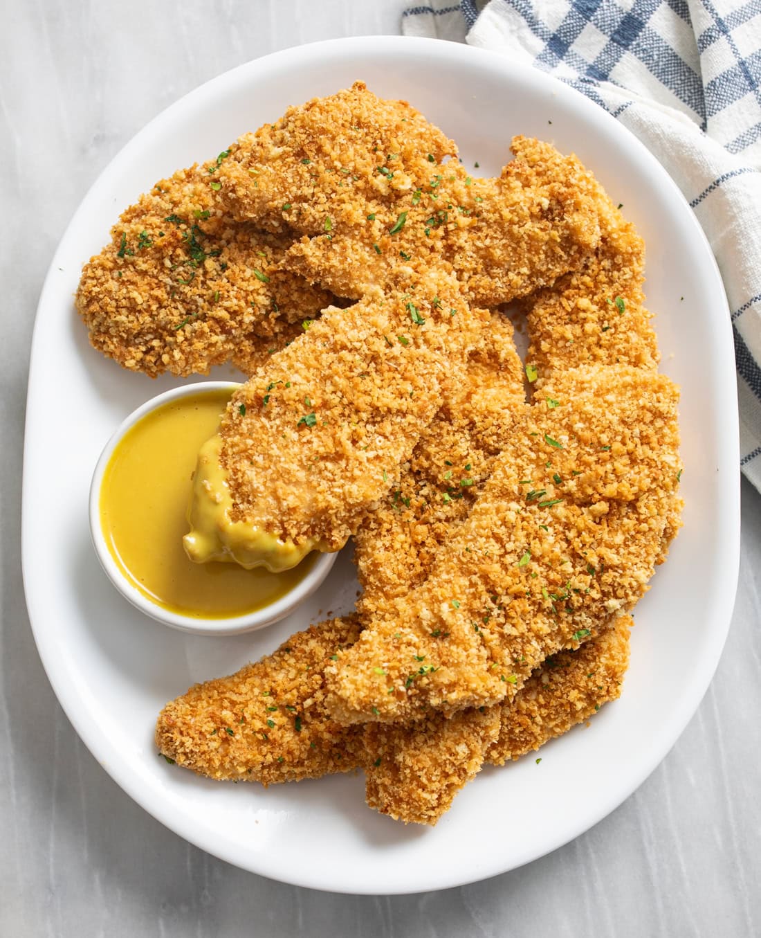 Overhead view of crispy Baked Chicken Tenders on a white platter with one being dipped into honey mustard sauce.