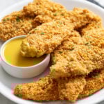 A white platter with crispy Baked Chicken Tenders stacked up with one dipped in honey mustard sauce.