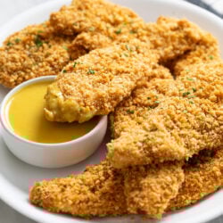 A white platter with crispy Baked Chicken Tenders stacked up with one dipped in honey mustard sauce.