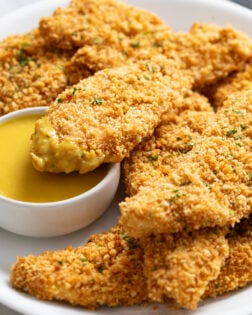 A white platter with crispy Baked Chicken Tenders stacked up with one dipped in honey mustard sauce.