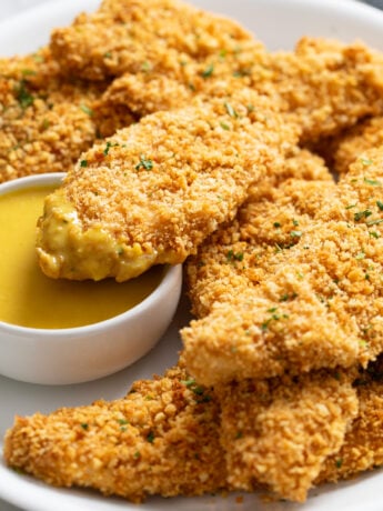 A white platter with crispy Baked Chicken Tenders stacked up with one dipped in honey mustard sauce.