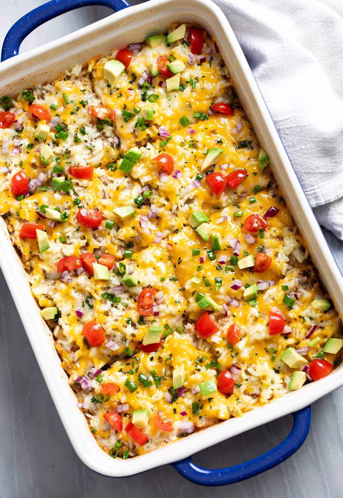 Burrito Casserole in a white and blue casserole dish with avocado, tomatoes, and onions on top.