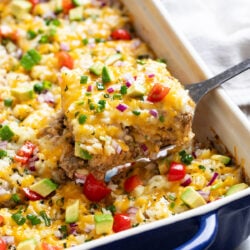 Burrito Casserole being scooped up from a casserole dish with a spatula. It's topped with rice and melted cheese with refried beans, tortillas, and ground beef underneath.