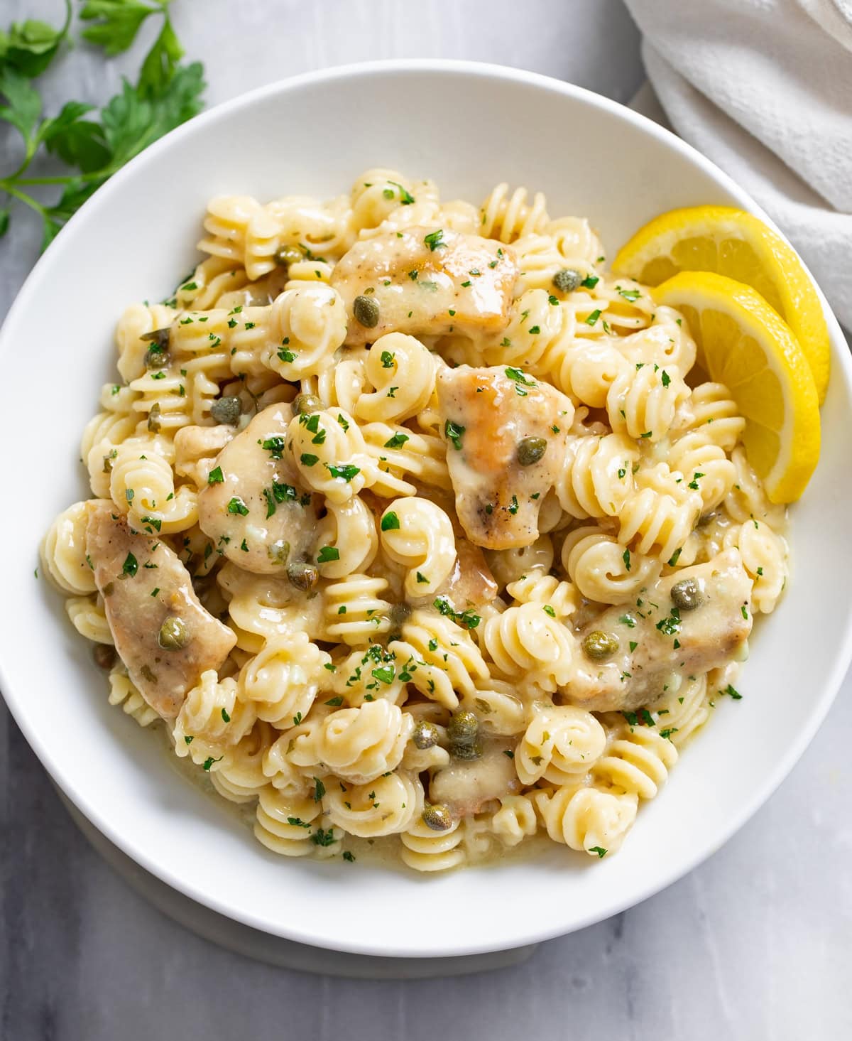 Chicken Piccata Pasta in a white bowl with chopped parsley on top and sliced lemon on the side.
