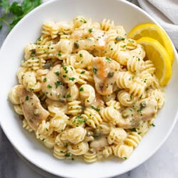 Chicken Piccata Pasta in a white bowl with a butter white wine piccata sauce, fresh parsley, and sliced lemon on the side.