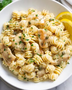 Chicken Piccata Pasta in a white bowl with a butter white wine piccata sauce, fresh parsley, and sliced lemon on the side.