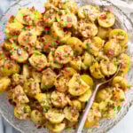 German Potato Salad in a glass bowl with a spoon on the side and bacon, chives, and dill on top.