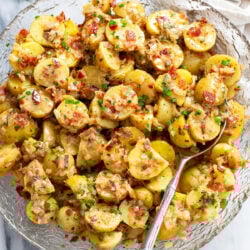 German Potato Salad in a glass bowl with a spoon on the side and bacon, chives, and dill on top.