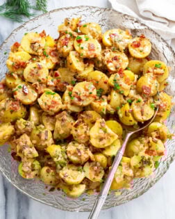 German Potato Salad in a glass bowl with a spoon on the side and bacon, chives, and dill on top.