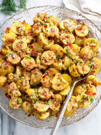 German Potato Salad in a glass bowl with a spoon on the side and bacon, chives, and dill on top.