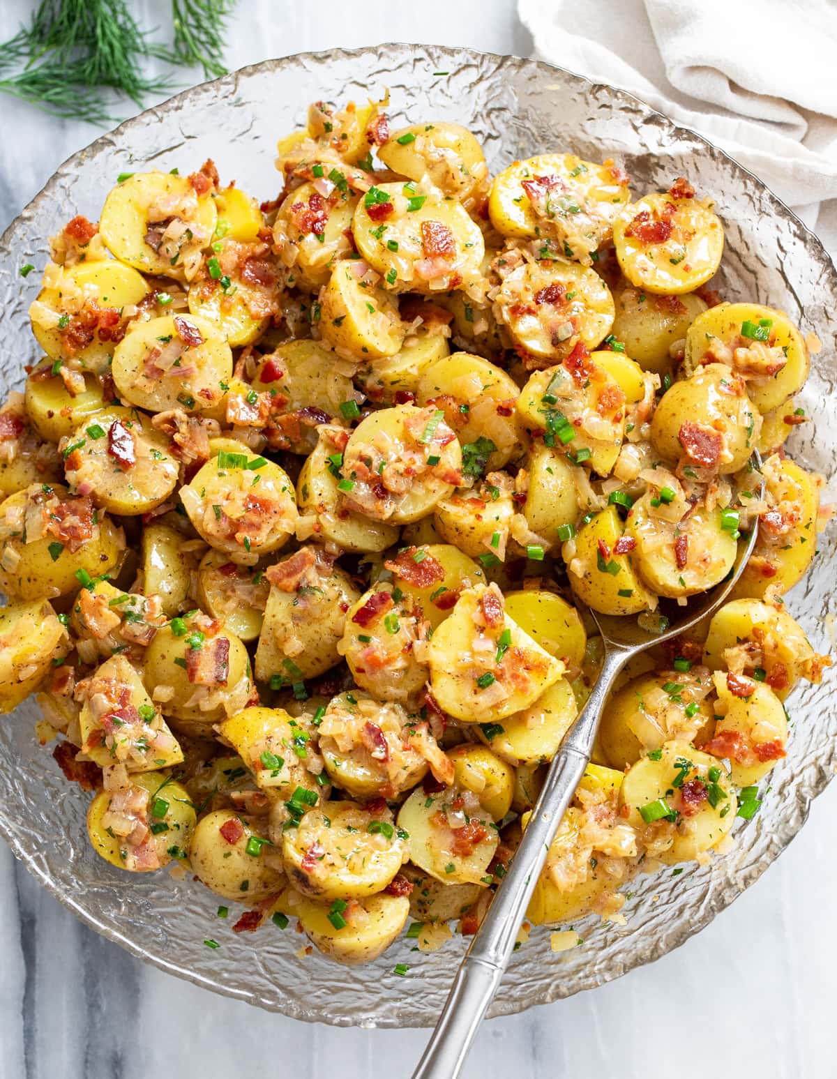 German Potato Salad in a glass bowl with a spoon on the side and bacon, chives, and dill on top.