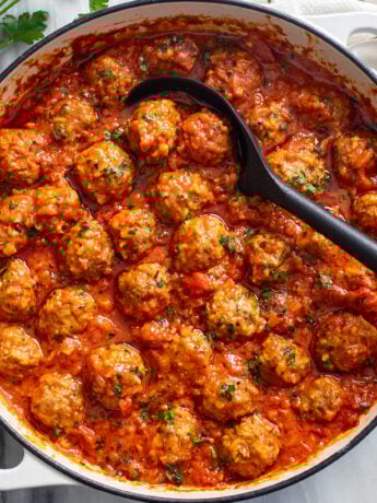 A white baking dish filled with Porcupine Meatballs in a red sauce with a spoon in the middle.