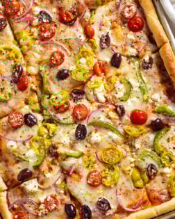 Greek Pizza topped with tomatoes, feta, peppers, olives, and pepperoncini peppers on a sheet pan.