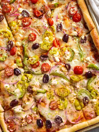 Greek Pizza topped with tomatoes, feta, peppers, olives, and pepperoncini peppers on a sheet pan.
