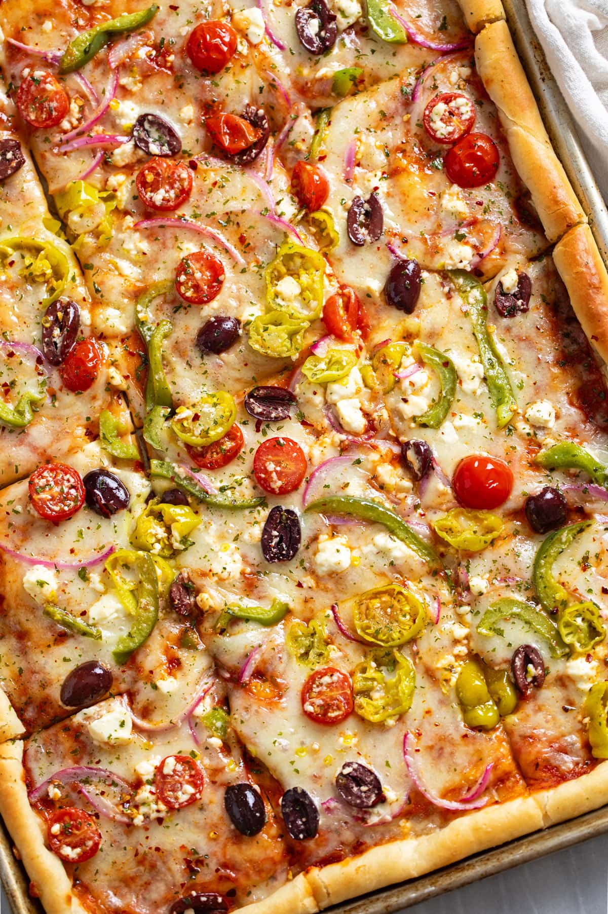 Greek Pizza topped with tomatoes, feta, peppers, olives, and pepperoncini peppers on a sheet pan.