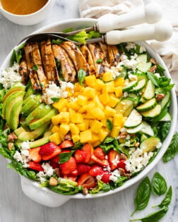 Mango Salad in a white bowl with chicken, sliced avocado, strawberries, cucumbers, mango, feta, and pecans.