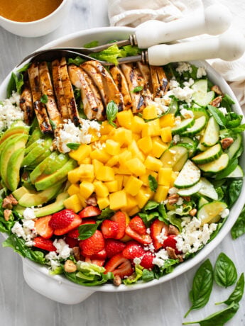 Mango Salad in a white bowl with chicken, sliced avocado, strawberries, cucumbers, mango, feta, and pecans.