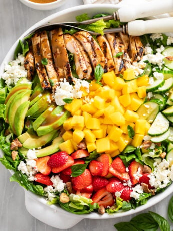 Mango Salad in a white bowl with grilled chicken, avocado, strawberry, and cucumber with mangos, feta, and dressing.