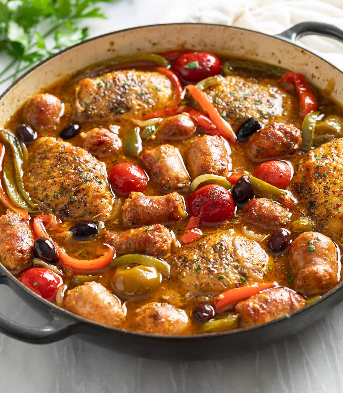 Side view of Chicken Scarpariello in a braiser with pan sauce, sausage, chicken thighs, peppers, and cherry peppers.