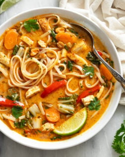 Thai Chicken Soup in a white bowl with noodles, vegetables, chicken, and a spoon and a lime wedge on the side.