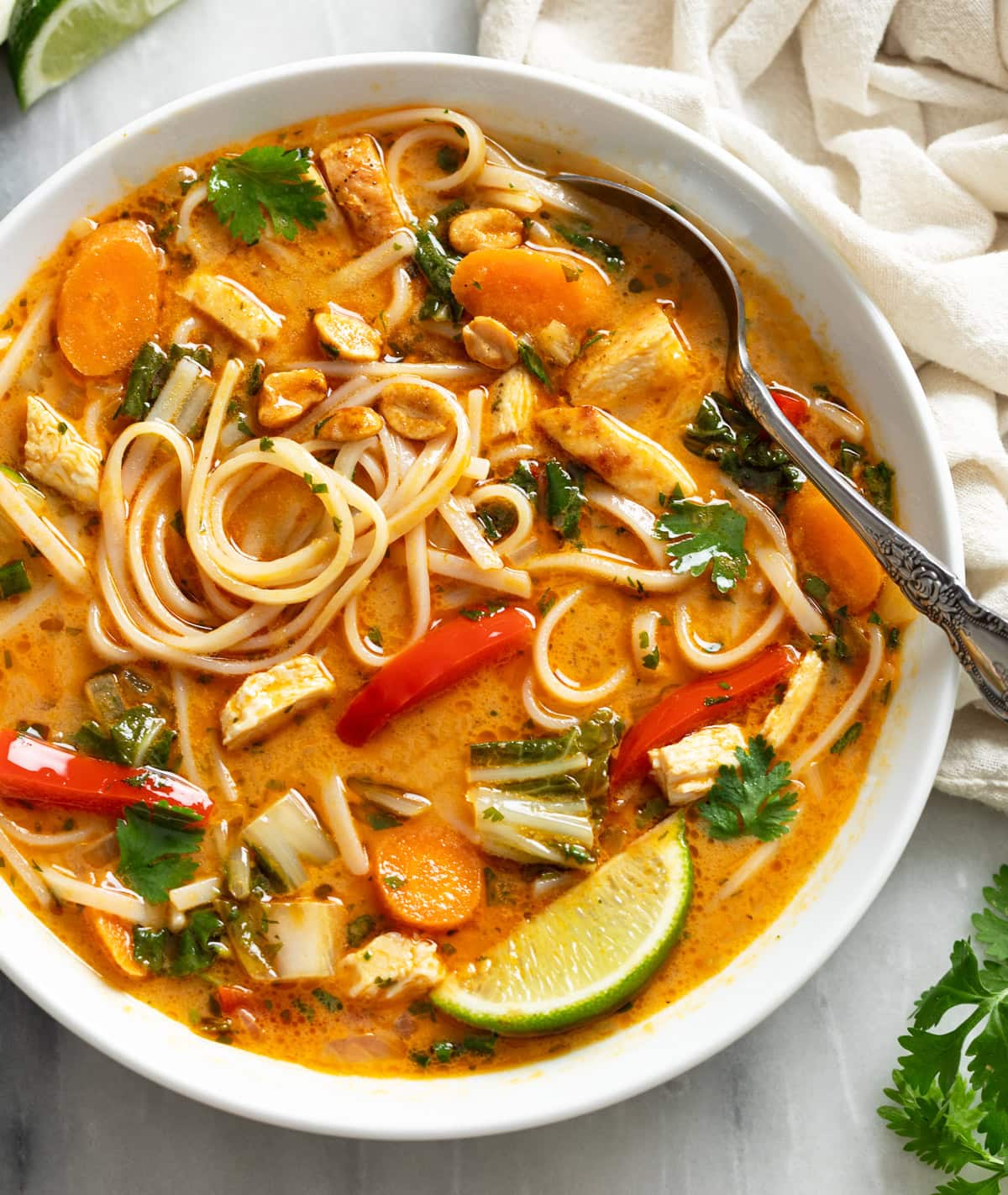 Thai Chicken Soup in a white bowl with noodles, vegetables, chicken, and a spoon and a lime wedge on the side.