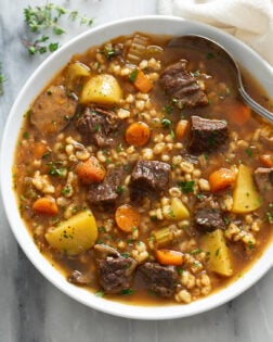 Beef Barley Soup in a white bowl with a spoon on the side and fresh parsley and thyme on top.