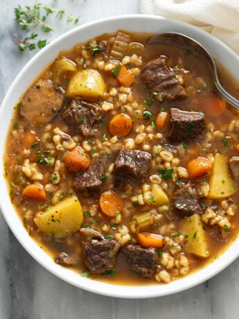 Beef Barley Soup in a white bowl with a spoon on the side and fresh parsley and thyme on top.