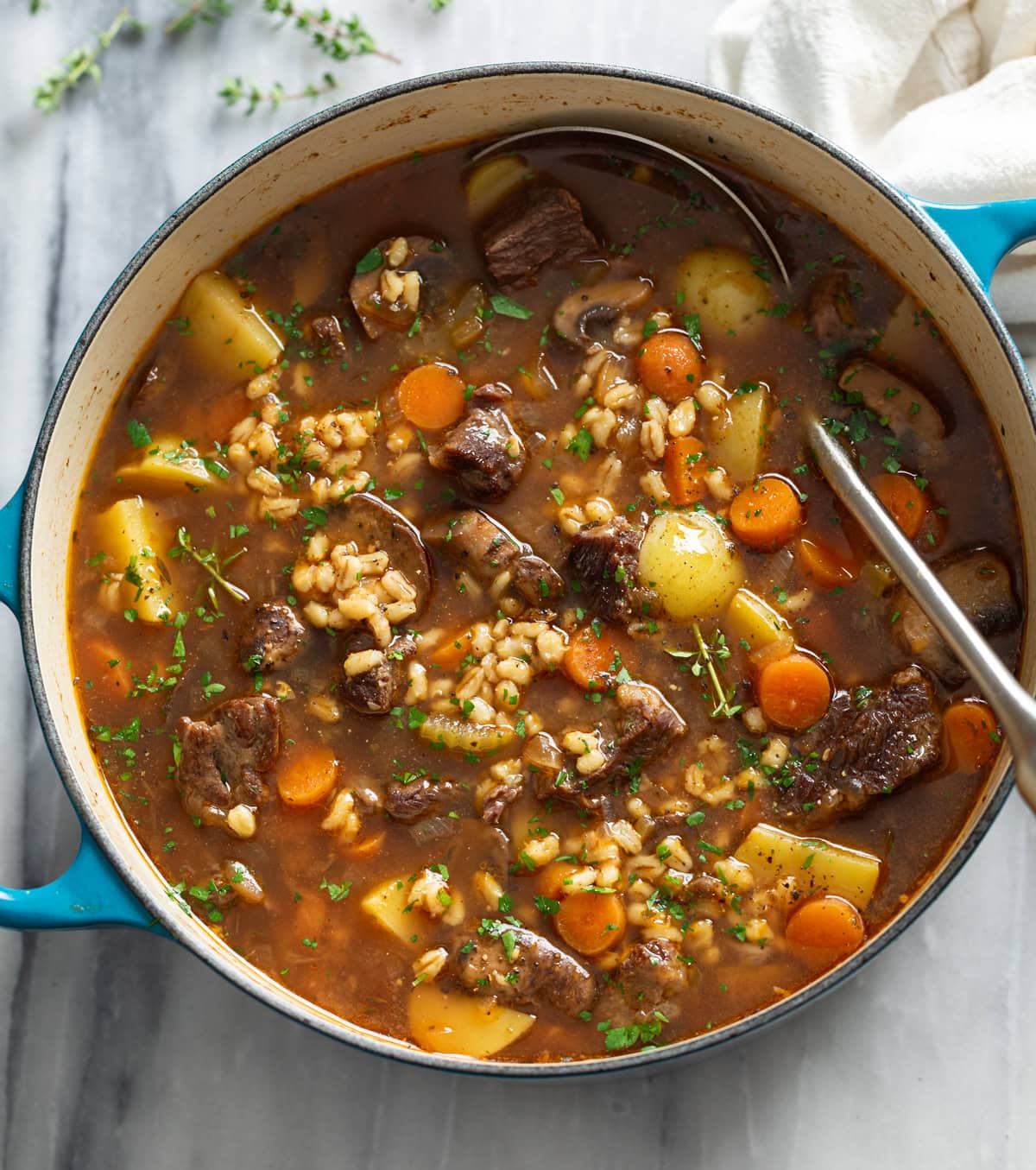 Beef Barley Soup in a soup pot with a ladle on the side and fresh parsley and thyme on top.