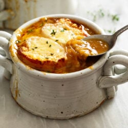 A crock of French Onion Soup with melted cheese and bread on top and a spoon scooping up some of the rich broth on the side.